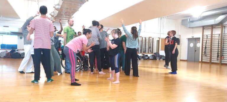 A diverse group of dancers, including performers with and without disabilities, engage in an inclusive movement exercise in a bright dance studio in Lisbon during the Biokinetics Workshop. Participants interact through expressive gestures and creative body dialogue.